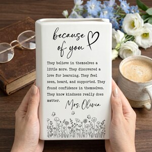 Puede incluir: Un florero rectangular de cer&aacute;mica blanca con texto negro que dice: because of you, seguido de un p&aacute;rrafo sobre la confianza y el aprendizaje, firmado Mrs. Olivia, con un dibujo lineal de flores silvestres y mariposas en la base.