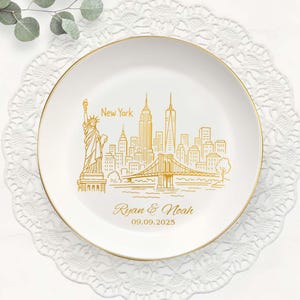 May include: A white ceramic plate with a gold rim, featuring a gold line drawing of the New York City skyline, including the Statue of Liberty and the Brooklyn Bridge. The plate is personalized with the names "Ryan & Noah" and the date "09.09.2025".