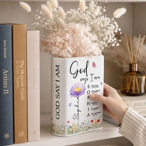 May include: A white rectangular ceramic vase featuring the text God says I Am, the name Sophia, a purple flower, and a colorful wildflower meadow print at the base. The vase includes the words Strong, Obedient, Healed, Innocent, and Accepted.
