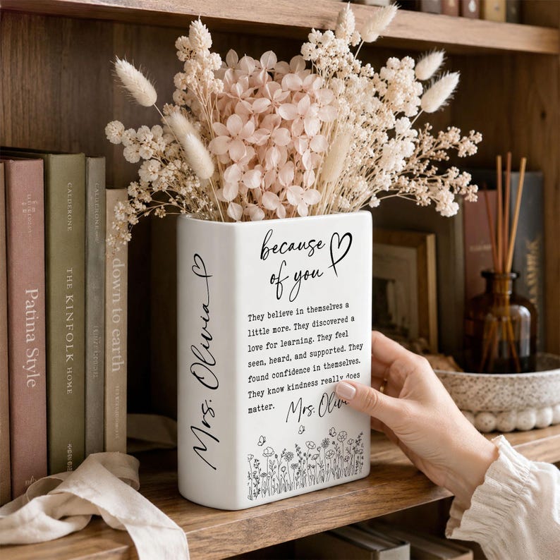 Puede incluir: Un jarr&oacute;n de cer&aacute;mica blanco con forma de libro que presenta texto negro que dice because of you y Mrs. Olivia, junto con un borde de arte lineal floral. El jarr&oacute;n est&aacute; lleno de flores secas beige y rosa claro, sobre un estante de madera.