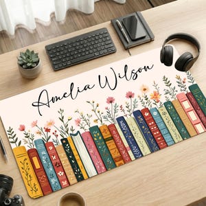 May include: A rectangular desk mat featuring a cream background with a row of colorful illustrated books at the bottom, topped with delicate wildflowers and the name Amelia Wilson written in black cursive script across the upper center.