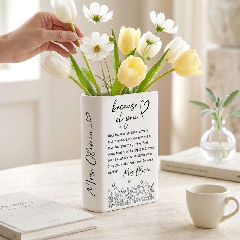 Puede incluir: Un florero de cer&aacute;mica blanca con forma de libro que presenta el texto 'because of you' y 'Mrs. Olivia' en letra cursiva negra. El florero contiene tulipanes blancos y amarillos y flores cosmos blancas, con una ilustraci&oacute;n floral en la base.