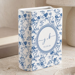 May include: A white ceramic book-shaped vase with a blue floral pattern. The vase features a circular design with the initials "j + k" and the date "02.20.26". The vase is a decorative item.