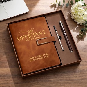 May include: A tan leather-effect journal with a metal clasp, engraved with the text "the OFFICIANT SCOTT WILSON" and "JACK & STEPHANIE DECEMBER 19, 2026". The journal is presented in a box alongside a dark brown pen and a silver metal pen refill.