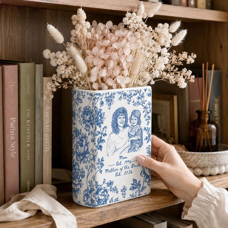 May include: A rectangular white ceramic vase featuring a blue floral pattern and a line art illustration of a person holding a child. The text reads Mom Est. 1996 and Mother of the Bride Est. 2026. The vase is filled with dried beige flowers.