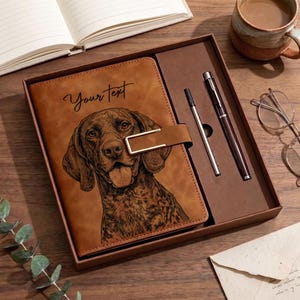 May include: A brown leather-look journal featuring a black line-art illustration of a dog and the text "Your text" on the cover. The journal includes a gold-toned metal clasp and is presented in a matching box with a silver-toned pen and a thin stylus.