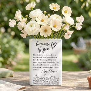 Puede incluir: Un jarr&oacute;n rectangular de cer&aacute;mica blanca lleno de flores blancas y crema. La parte frontal presenta texto negro que dice "because of you" con un coraz&oacute;n, seguido de un p&aacute;rrafo sobre confianza y bondad, firmado "Mrs. Olivia" sobre un borde floral.