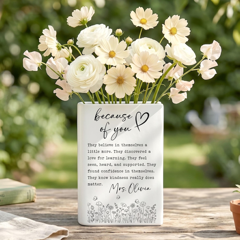 Puede incluir: Un jarr&oacute;n rectangular de cer&aacute;mica blanca lleno de flores blancas y crema. La parte frontal presenta texto negro que dice "because of you" con un coraz&oacute;n, seguido de un p&aacute;rrafo sobre confianza y bondad, firmado "Mrs. Olivia" sobre un borde floral.