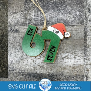 Peut inclure: Ornement de Noël en bois avec les chiffres "6" et "7" en vert, avec les mots "SIX" et "SEVEN" sur chaque chiffre. Un bonnet de Père Noël avec le mot "DOM" et le chiffre "25" est au sommet. L'ornement est suspendu à une ficelle.
