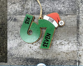 May include: Wooden Christmas ornament featuring the numbers "6" and "7" in green with the word "SIX" and "SEVEN" on each number. A Santa hat with the word "DOM" and the number "25" is on top. The ornament hangs from a twine string.