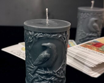 May include: A gray candle with a detailed design of a raven and foliage. The candle is sitting on a surface with a deck of tarot cards.