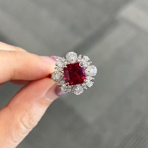 Peut inclure: Gros plan sur une bague ornée d'une grosse pierre rouge carrée, entourée de petites pierres claires. Le sertissage de la bague semble être en or ou en métal doré, avec les petites pierres disposées en forme de fleur ou de halo.