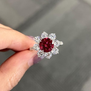 Peut inclure: Une bague en forme de fleur avec une grosse pierre rouge foncé ronde au centre, entourée de petites pierres claires. Le sertissage est complexe et le métal est argenté. La bague est présentée sur un fond flou.