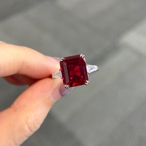 Puede incluir: Un anillo de plata con una gran gema roja oscura rectangular como pieza central. Dos piedras rectangulares claras más pequeñas flanquean la piedra principal. El anillo se sostiene sobre un fondo borroso.