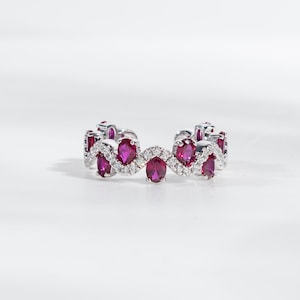 Może przedstawiać: Srebrny pierścionek z powtarzającym się wzorem owalnych, głęboko czerwonych kamieni szlachetnych, przeplatanych mniejszymi, okrągłymi, przezroczystymi kamieniami szlachetnymi. Pierścionek jest umieszczony na białym tle, podkreślając blask kamieni.