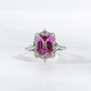 Op de afbeelding: Een zilveren ring met een grote, rechthoekige, roze edelsteen, omringd door een halo van kleinere, heldere edelstenen. De ringband is ook versierd met kleine, heldere edelstenen, wat een sprankelend en elegant ontwerp creëert. Op een witte achtergrond.