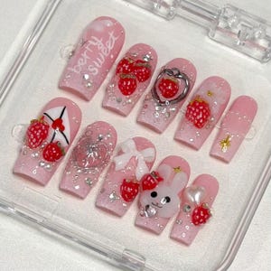May include: Pink artificial nails with strawberry and bunny designs. Some nails feature the text "Berry Sweet". Embellishments include rhinestones, bows, and heart-shaped charms. The nails are in a clear plastic case.