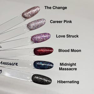 May include: Six clear plastic nail polish display sticks with oval tips painted in glittery shades. The colors are labeled as The Change, Career Pink, Love Struck, Blood Moon, Midnight Massacre, and Hibernating, featuring pink, purple, red, blue, and black tones.