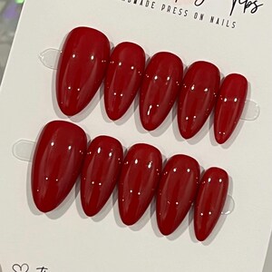 May include: Two sets of red press-on nails in various sizes. The nails have a glossy finish and an oval shape. The nails are arranged on a white card with the text "HANDMADE PRESS ON NAILS".
