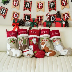 May include: Five Christmas stockings with embroidered names and woodland animal designs, including a polar bear, squirrel, owl, and fox. The stockings are red and beige with white faux fur trim. A banner that says "Happy Holiday" hangs above.