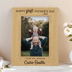 May include: A light-coloured wooden picture frame with the text "Happy First Father's Day 2025" at the top. The frame holds a photo of a man with a child on his shoulders. The text "I Love You, Daddy" and a name are at the bottom.