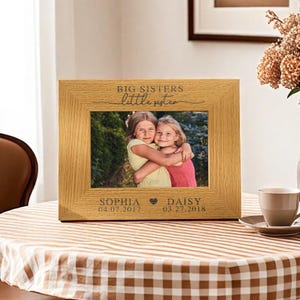 Puede incluir: Un marco de fotos de madera con una foto de dos hermanas abrazándose. El marco está grabado con el texto "Big Sisters little sister" y los nombres "Sophia" y "Daisy" con sus fechas de nacimiento.