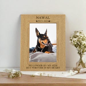 May include: A wooden photo frame with a picture of a dog inside. The frame has the words "NAWAL 2015-2025" and "NO LONGER BY MY SIDE BUT FOREVER IN MY HEART" engraved on it.
