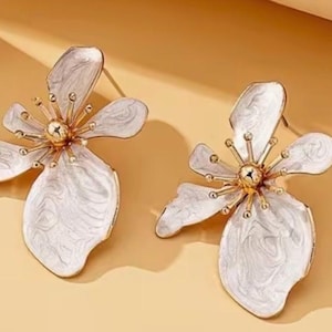 May include: A pair of white and gold flower-shaped earrings. The earrings have a gold center with white petals and a gold outline. The earrings are on a light orange background.