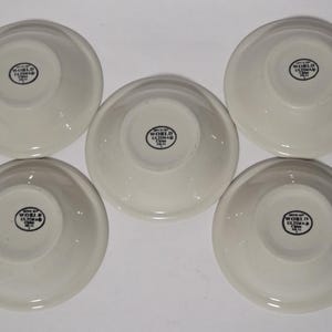 May include: A collection of six off-white ceramic bowls, each with a rounded shape and a slightly raised rim. The bowls are arranged in a circular pattern. Each bowl has a black circular stamp that reads "WORLD ULTIMA" and "CHINA" in the center.