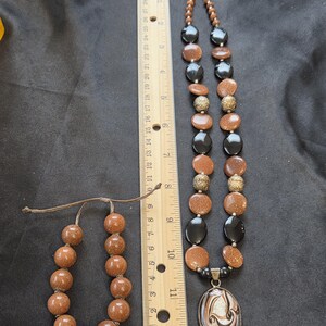 May include: A jewelry set featuring a necklace, bracelet, and pendant. The necklace has alternating black, brown, and goldstone beads, with a silver pendant. The bracelet is made of brown beads. A ruler shows the necklace is approximately 11 inches long.