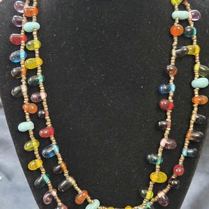 May include: A colorful beaded necklace with teardrop-shaped beads in various colors, including red, blue, yellow, and turquoise. The beads are strung on a gold-colored chain, creating a layered effect. The necklace is displayed on a black surface.