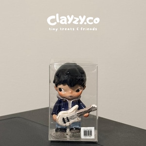 May include: A small figurine of a person with dark hair wearing a blue jacket and jeans, holding a white electric guitar. The figure is encased in a clear plastic box with the text clayzy.co tiny treats & friends printed above it.