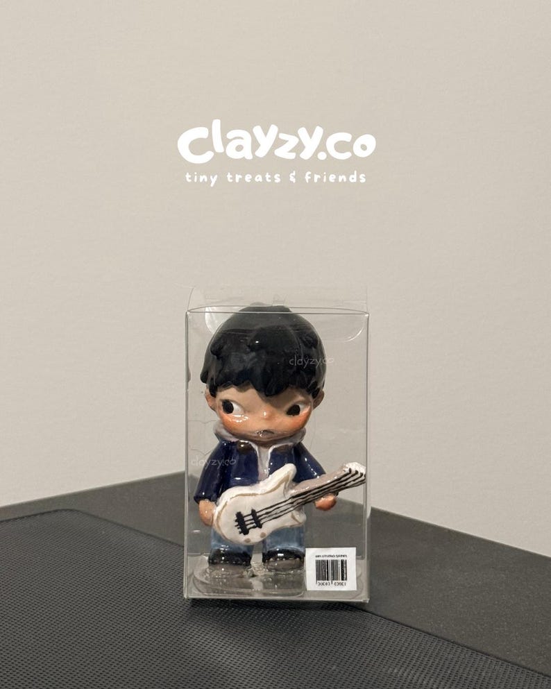 May include: A small figurine of a person with dark hair wearing a blue jacket and jeans, holding a white electric guitar. The figure is encased in a clear plastic box with the text clayzy.co tiny treats & friends printed above it.