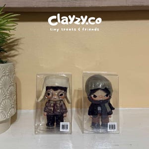 May include: Two small figurines in clear plastic boxes. The left figurine wears a cream-colored trapper hat, round glasses, and a brown patterned jacket. The right figurine wears a grey beanie, a dark scarf, and a black jacket. Text reads clayzy.co.