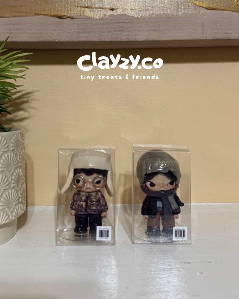 May include: Two small figurines in clear plastic boxes. The left figurine wears a cream-colored trapper hat, round glasses, and a brown patterned jacket. The right figurine wears a grey beanie, a dark scarf, and a black jacket. Text reads clayzy.co.