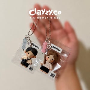 Puede incluir: Dos figuras de arcilla en miniatura de un hombre y una mujer, cada una en una bolsa de plástico transparente con un llavero. La figura del hombre lleva pantalones cortos negros y una camiseta negra. La figura de la mujer lleva una camiseta blanca y pantalones cortos negros. La marca "Clayzy.co" es visible.
