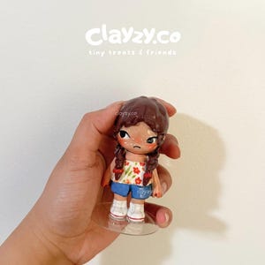 May include: A small figurine with brown hair in two braids, wearing a white tube top with a red and green floral pattern, blue shorts, and white sneakers. The figurine has a sad facial expression with large dark eyes and stands on a clear base.