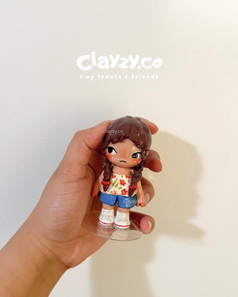 May include: A small figurine with brown hair in two braids, wearing a white tube top with a red and green floral pattern, blue shorts, and white sneakers. The figurine has a sad facial expression with large dark eyes and stands on a clear base.