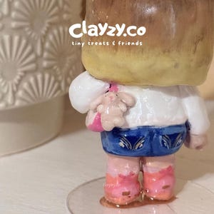 May include: A small figurine with brown hair, a white top, blue shorts, and pink shoes, featuring a small pink bag with a tiny rabbit charm. The text clayzy.co tiny treats & friends is visible on the upper portion of the figurine's head.