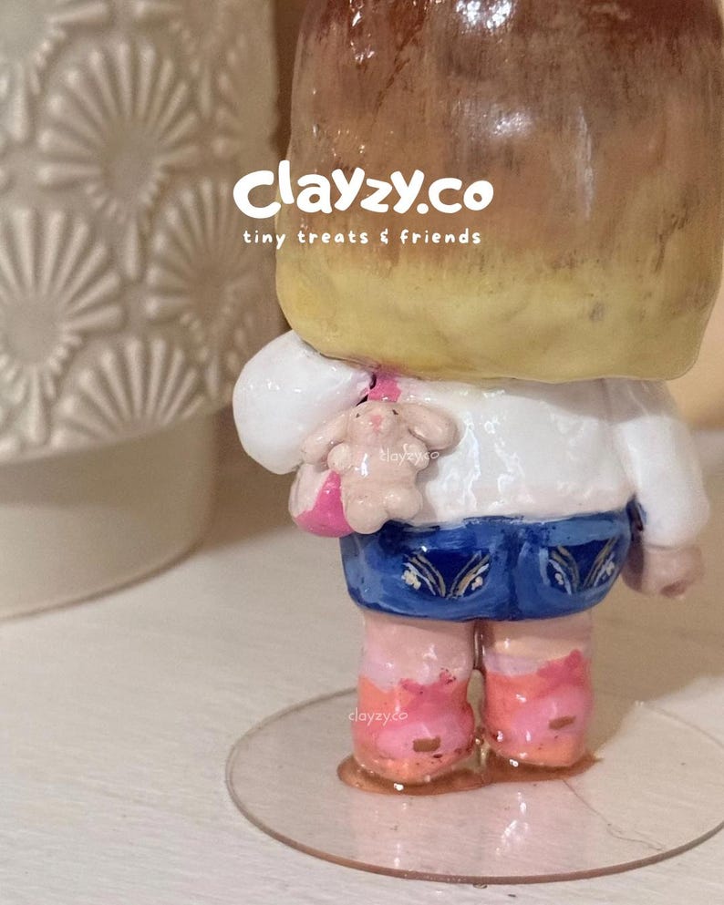 May include: A small figurine with brown hair, a white top, blue shorts, and pink shoes, featuring a small pink bag with a tiny rabbit charm. The text clayzy.co tiny treats & friends is visible on the upper portion of the figurine's head.