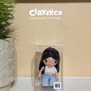 May include: A small figurine of a person with long black hair, wearing a white crop top and light blue jeans, encased in a clear plastic box. The text clayzy.co tiny treats & friends is visible above the figurine, and a barcode label is on the box.