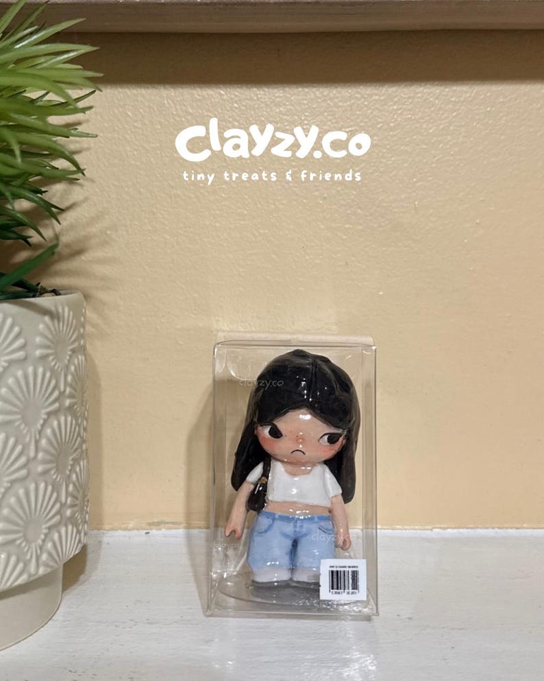 May include: A small figurine of a person with long black hair, wearing a white crop top and light blue jeans, encased in a clear plastic box. The text clayzy.co tiny treats & friends is visible above the figurine, and a barcode label is on the box.