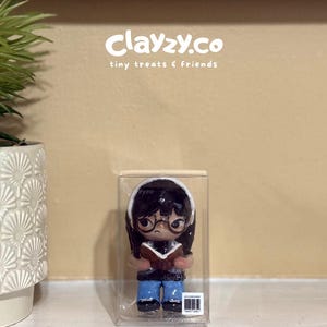 May include: A small figurine of a character with dark hair, glasses, and a white headband, wearing a dark top and blue pants while holding an open book. The figurine is encased in a clear rectangular plastic box with the text clayzy.co tiny treats & friends.