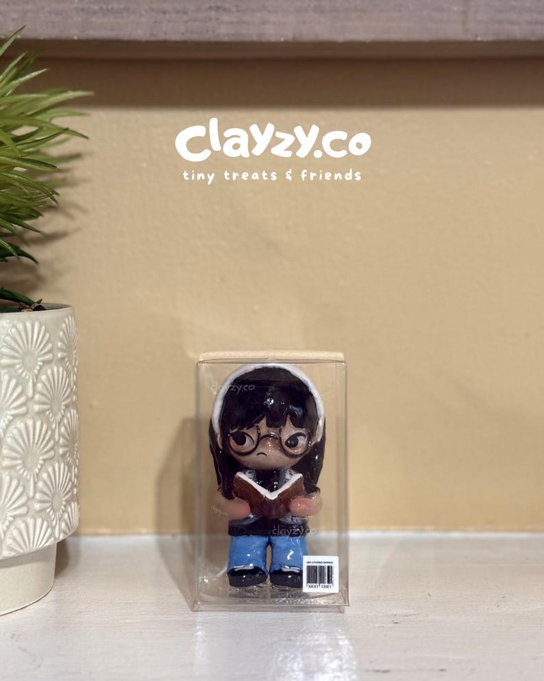 May include: A small figurine of a character with dark hair, glasses, and a white headband, wearing a dark top and blue pants while holding an open book. The figurine is encased in a clear rectangular plastic box with the text clayzy.co tiny treats & friends.