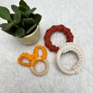 Op de afbeelding: Drie handgemaakte macramé bijtringen voor baby's in oranje, roest en crèmekleur. Elke bijtring heeft een houten ringbasis. Op de achtergrond staat een kleine vetplant in een pot.
