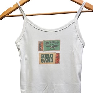 May include: A white ribbed tank top featuring a graphic print of two matchboxes. The top matchbox is green with an eagle and the text GO BIRDS. The bottom matchbox is tan with the text BIRD GANG in green letters and red match tips visible.