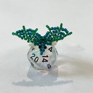 May include: A clear iridescent twenty-sided die with black numbers, 4, 6, 8, 10, 12, 14, 16, 18, 20, and a green beaded dragon perched on top.