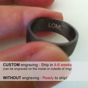 May include: A black ring with the inscription "LOML" engraved on the inside. The ring is being held by a finger.