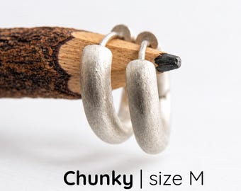 Könnte beinhalten: Ein Paar klobige Creolen aus massivem 925er Sterlingsilber mit matter Oberfläche, die auf der Spitze eines Holzbleistifts ruhen. Der Text darunter lautet: Chunky | size M solid 925 sterling silver.