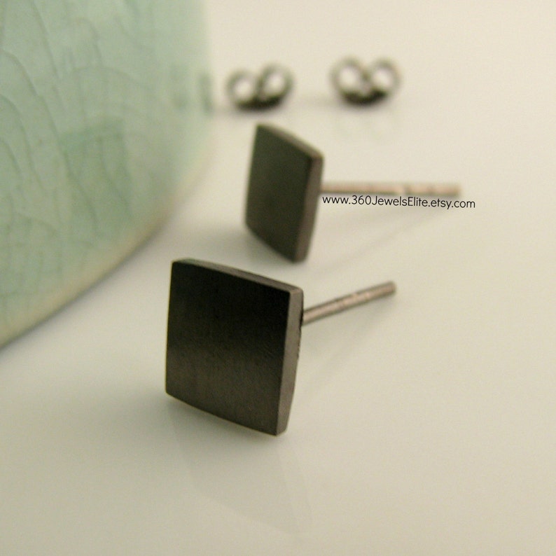 May include: A pair of black square stud earrings. The earrings are made of metal and have a brushed finish. The earrings are shown on a white surface.